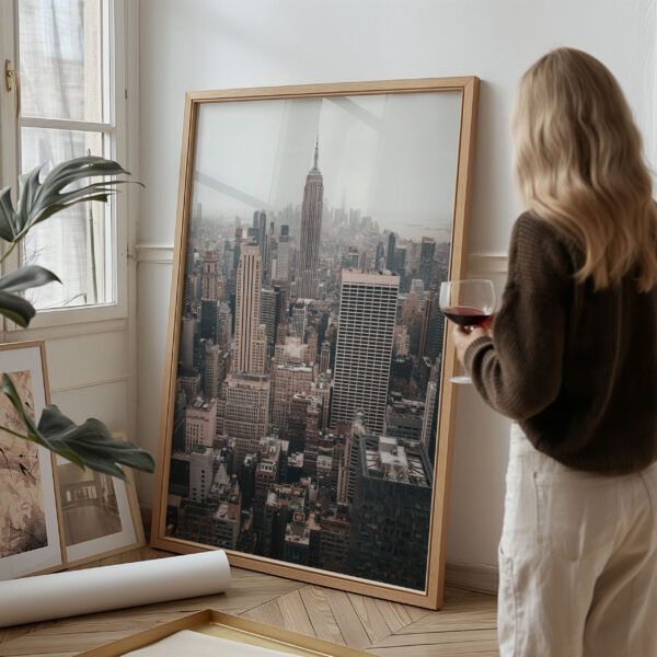 Large framed print of the NYC skyline featuring the Empire State Building, leaning against a wall beside a woman in a cozy home setting.