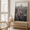 Neutral-toned interior with a framed Empire State Building skyline print leaning on a wooden bench near a window and minimalist ceramic vase.