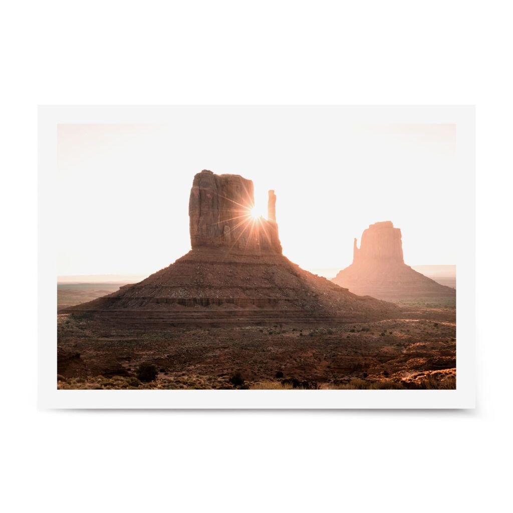Sunlight flaring through the iconic butte formations in Monument Valley at sunrise, casting warm light over the desert landscape.