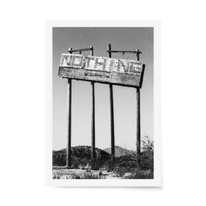 Black and white photograph of a vintage roadside "Nothing" sign standing in the Arizona desert with scrubland and distant mountains.
